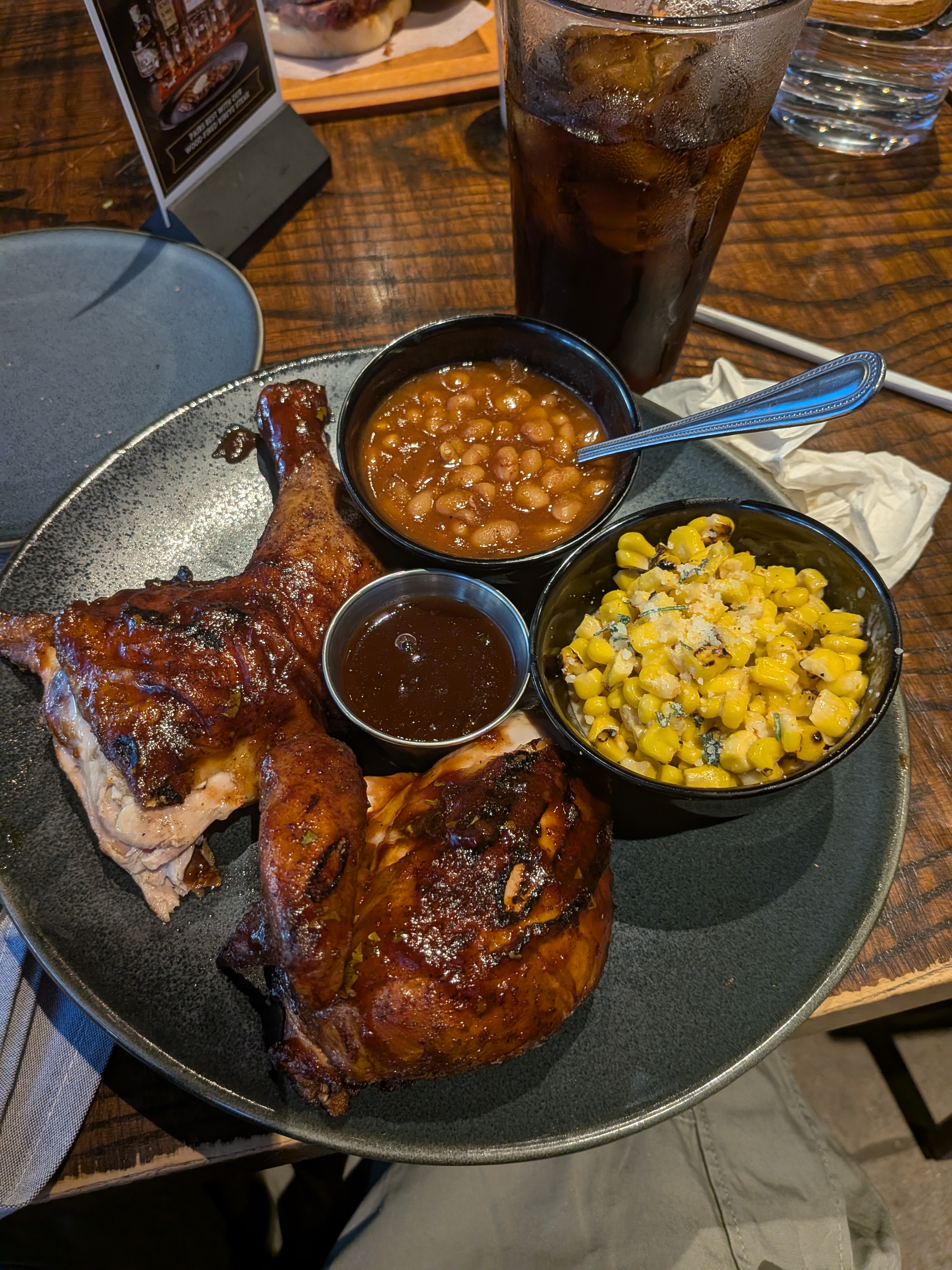 A plate of BBQ chicken with sides of baked beans and corn and a cup of BBQ sauce