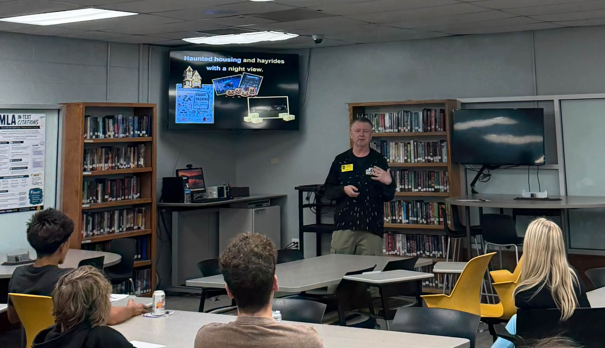 Charles speaking to a class of students in the high school library, with a slide on the screen behind him