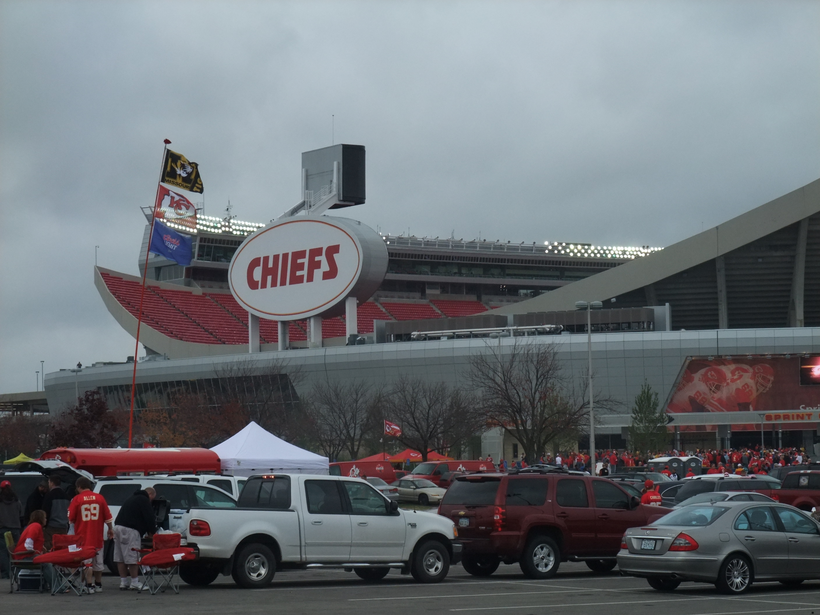 Arrowhead Stadium on a game day, with the Chiefs sign, tailgating tents, and cars filling the lot under a grey sky