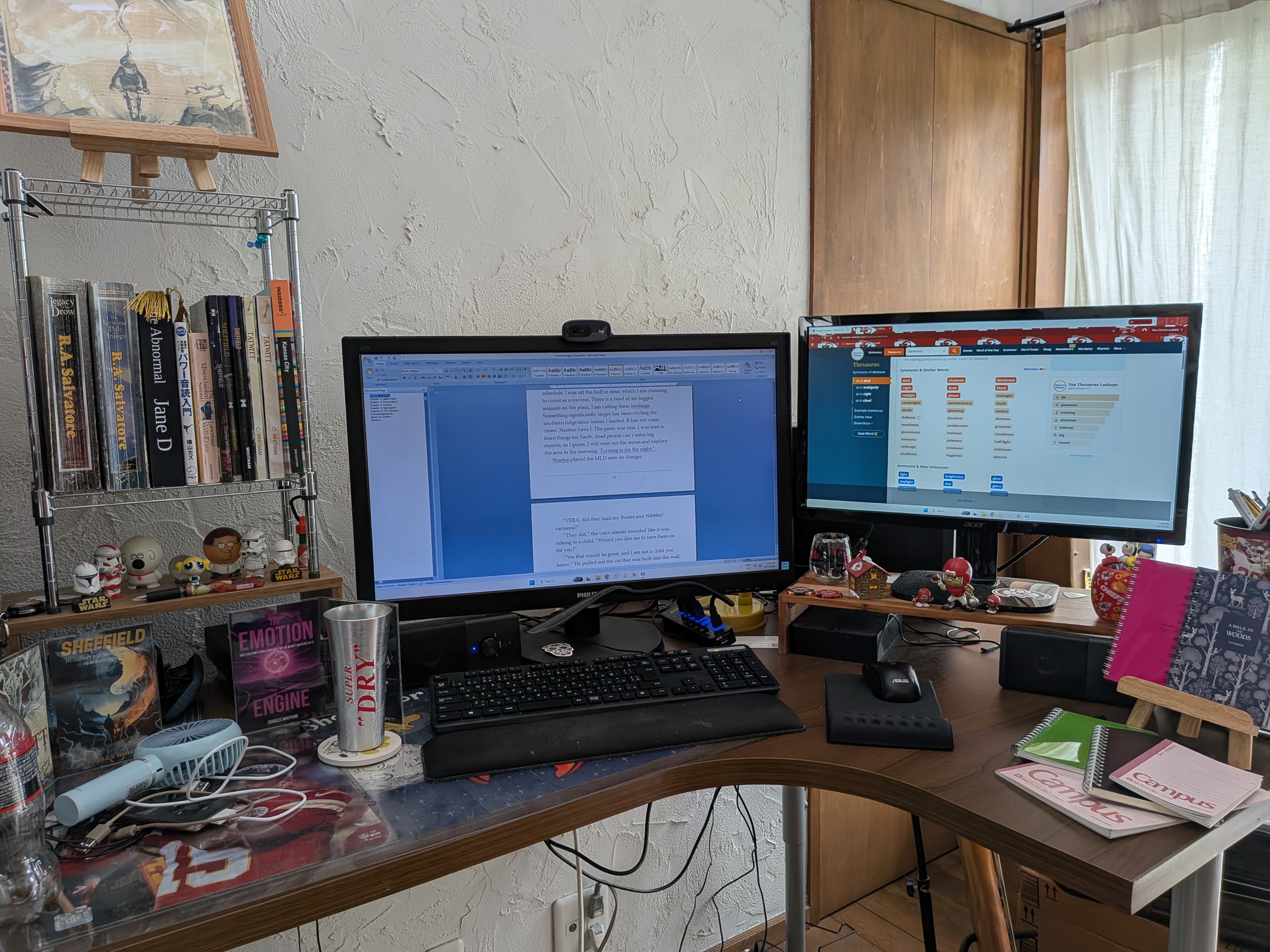 The writing desk setup, with dual monitors, the manuscript open, and books on the shelf beside it